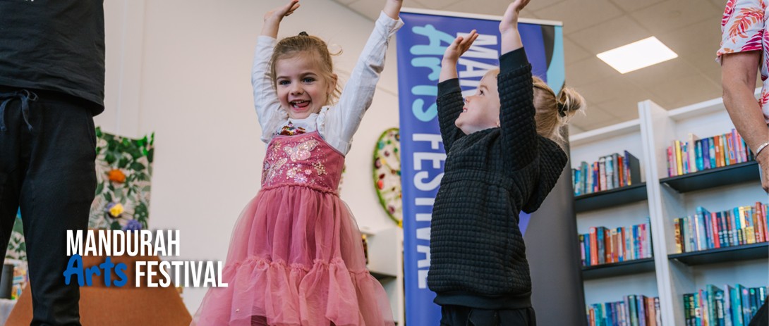 Two young children lift their hands up in joy and smile at the camera in a library setting